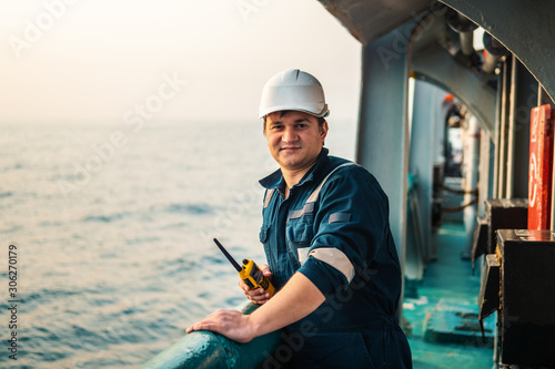 Marine Deck Officer or Chief mate on deck of offshore vessel or ship , wearing PPE personal protective equipment - helmet, coverall. He holds VHF walkie-talkie radio in hands. photo