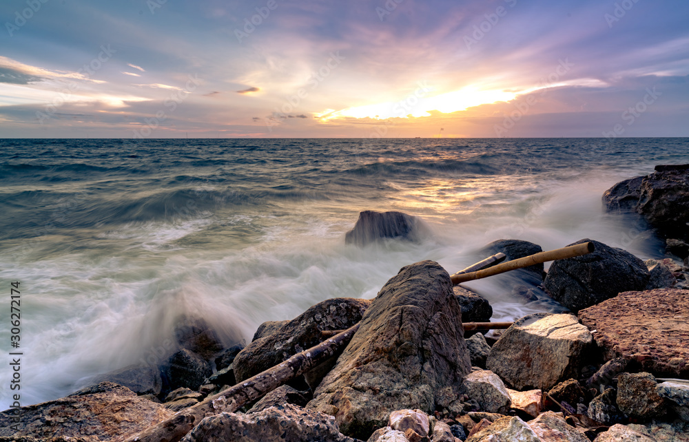 Ocean water splash on rock beach with beautiful sunset sky and clouds. Sea wave splashing on stone at sea shore on summer. Nature landscape. Tropical paradise beach at sunset. Rock beach at coast.