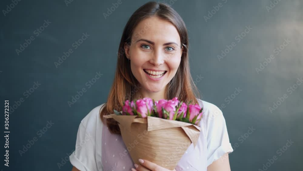 Slow motion of pretty young lady holding bunch of flowers smelling smiling enjoying pink roses standing on gray background. People, gifts and dates concept. Slow motion of pretty young lady holding bunch of flowers smelling smiling enjoying pink roses standing on gray background. People, gifts and dates concept.