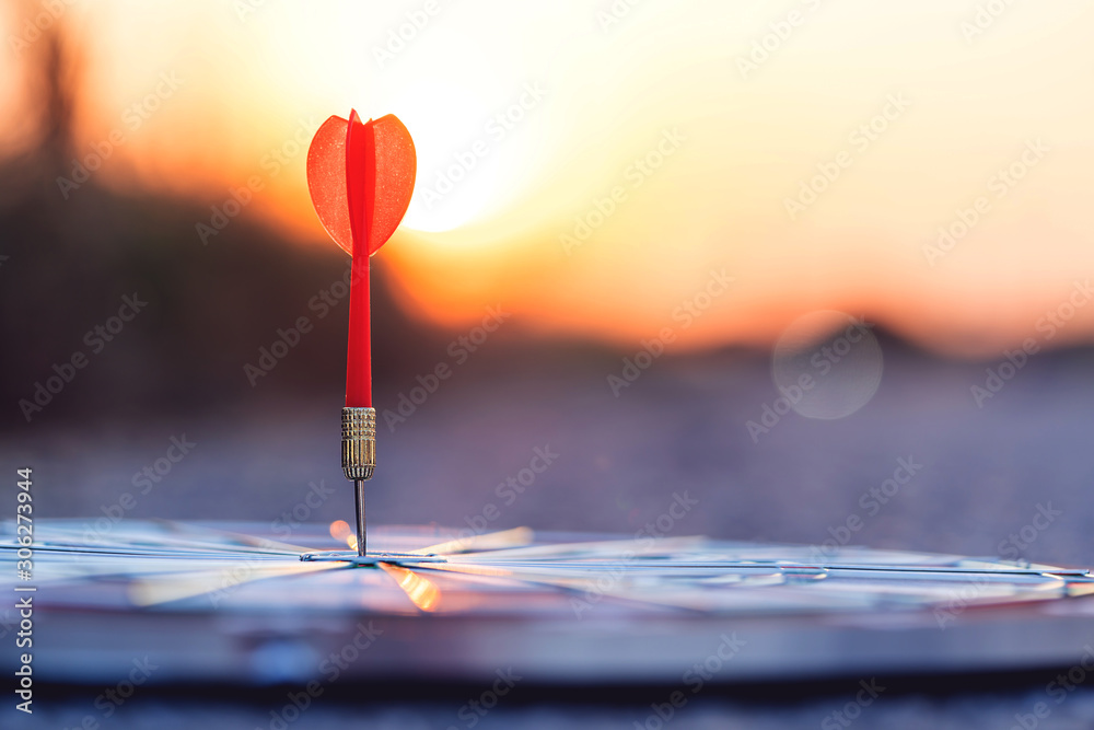 Close up red dart arrow hitting target center dartboard on sunset background Close up red dart arrow hitting target center dartboard on sunset background