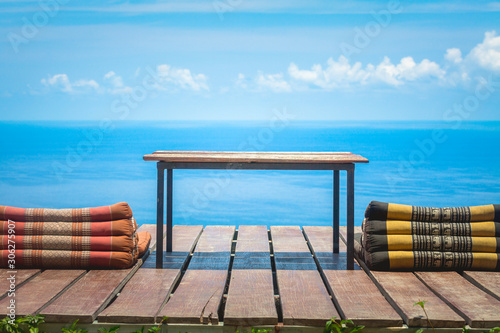 Fototapeta Naklejka Na Ścianę i Meble -  Table and Thai Triangle Pillows on Wooden Terrace with Tropical Beach, Blue Sky and Clouds in Background