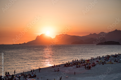 Orange sunset by the ocean in Piratininga, Niteroi, with sun dipping behing the Gavea Stone in Rio de Janeiro. A beach full of people can be seen in the foreground in shadow. photo