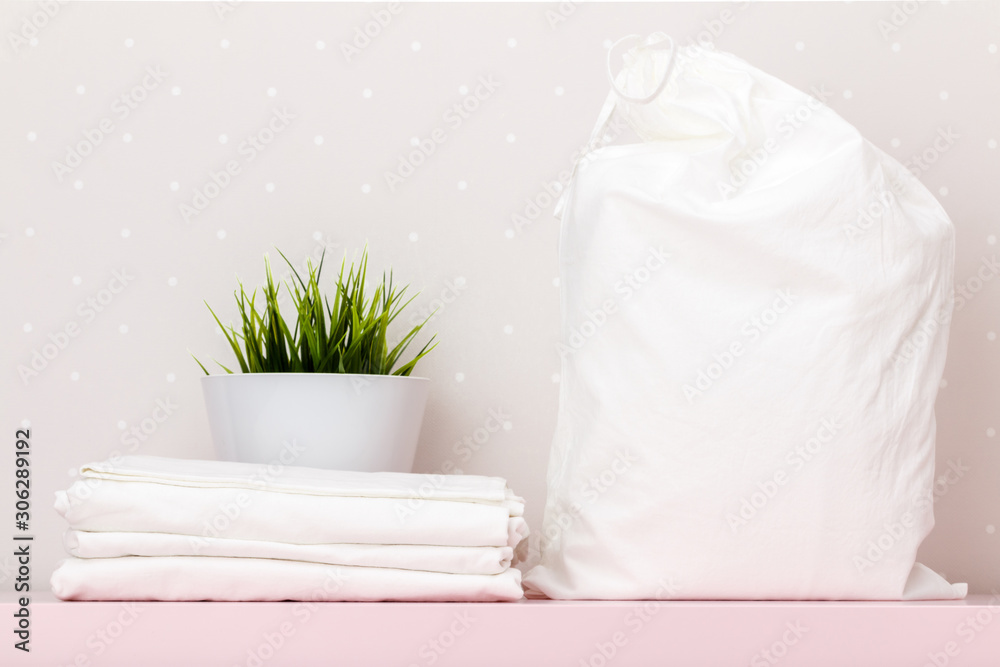 Close-up of a stack of white bedding, storage bag, house plant on a table against the background of light wall Close-up of a stack of white bedding, storage bag, house plant on a table against the background of light wall