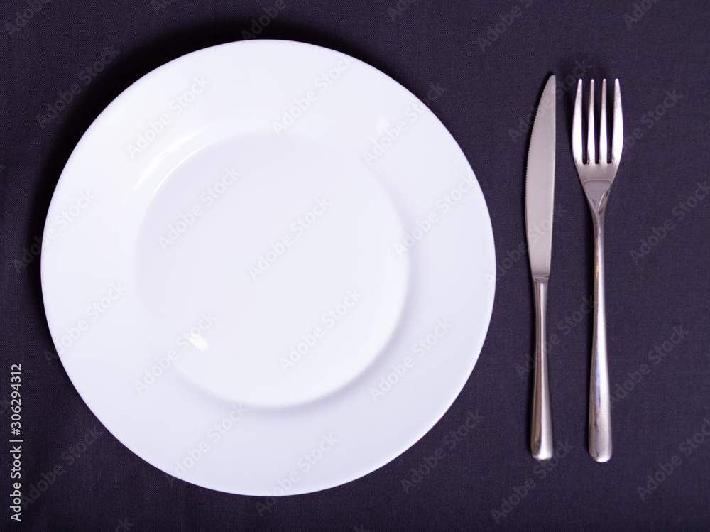white plate, knife and fork on napkin on dark tablecloth white plate, knife and fork on napkin on dark tablecloth