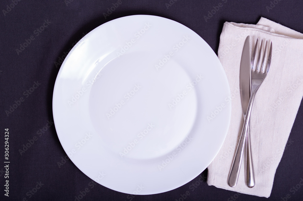 white plate, knife and fork on napkin on dark tablecloth white plate, knife and fork on napkin on dark tablecloth