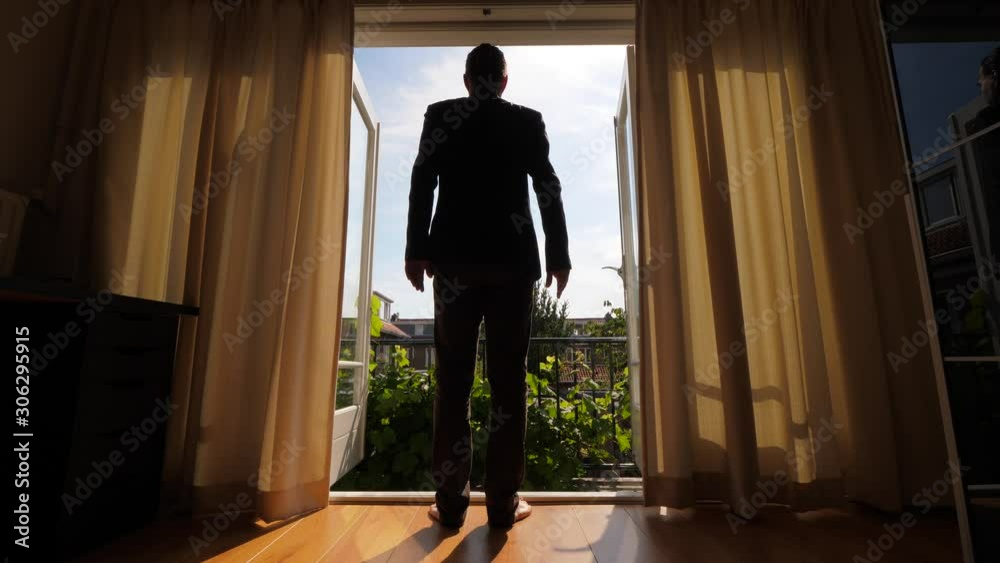 Man walk in room and open glass leaves of balcony door, stay with arms on hips and looking out to inner yard of the house. Sunny summer weather, late morning time on noon, cozy bedroom at second floor Man walk in room and open glass leaves of balcony door, stay with arms on hips and looking out to inner yard of the house. Sunny summer weather, late morning time on noon, cozy bedroom at second floor