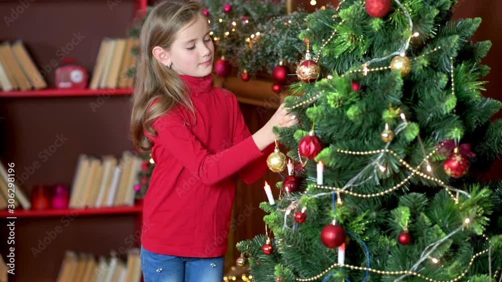 Cute girl in a red sweater decorates the Christmas tree in the room with decorative balls. Preparing for Christmas and New Year. Holidays Cute girl in a red sweater decorates the Christmas tree in the room with decorative balls. Preparing for Christmas and New Year. Holidays