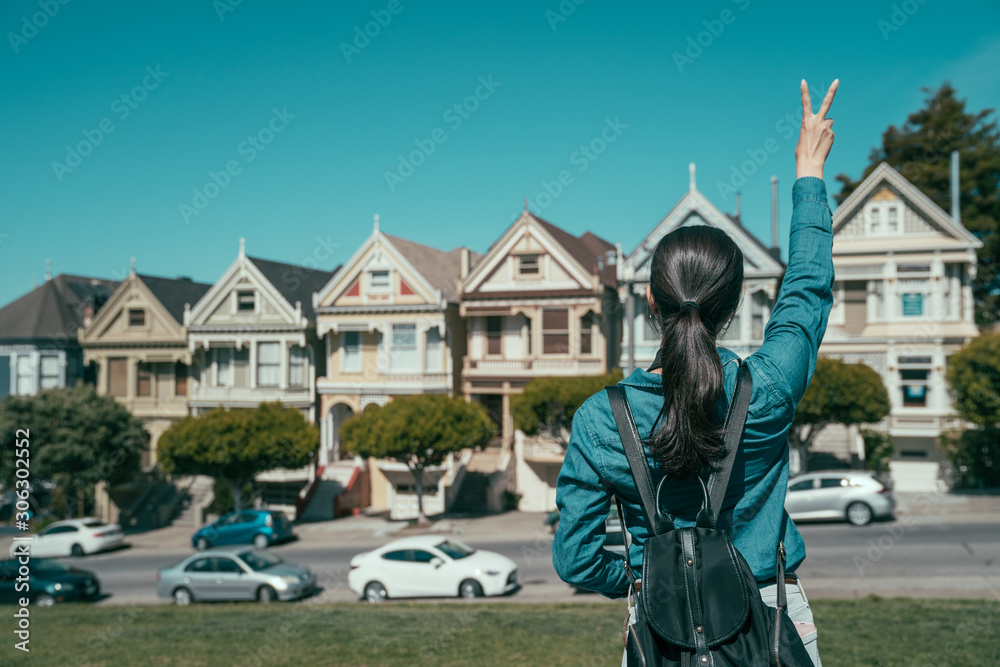 back view portrait of young girl with backpack showing v sign while raising hands. female tourist sightseeing in san francisco with famous attraction of painted ladies on bright sunny day.