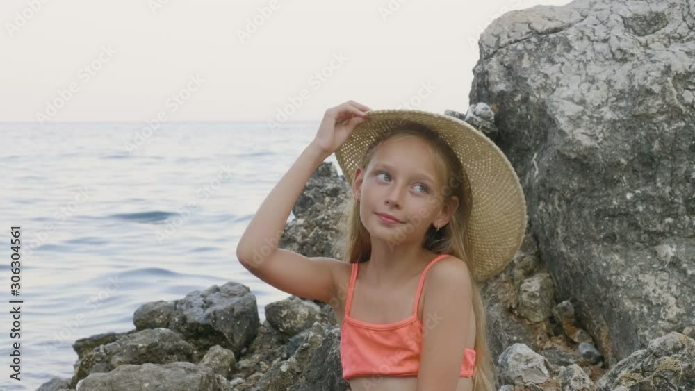 Cute teenager girl in bikini trying straw hat on stone in sea ...