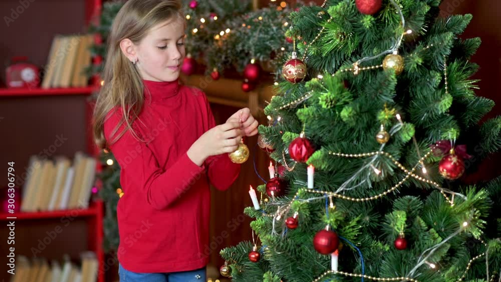 Cute girl in a red sweater decorates the Christmas tree in the room with decorative balls. Preparing for Christmas and New Year. Holidays Cute girl in a red sweater decorates the Christmas tree in the room with decorative balls. Preparing for Christmas and New Year. Holidays