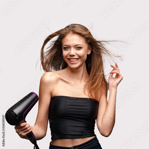A young attractive girl is blow-drying her hair and laughing.