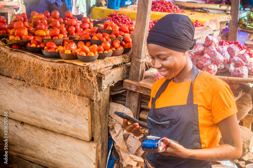 young african woman selling in a local african market holding a mobile point of sale system and using her mobile phone