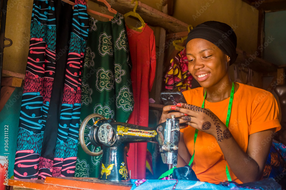 young african woman who is a tailor smiling while using her mobile phone - obrazy, fototapety, plakaty 