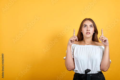 Scared young woman pointing up, afraid, looking shocked isolated on orange background in studio in casual white shirt. People sincere emotions, lifestyle concept.