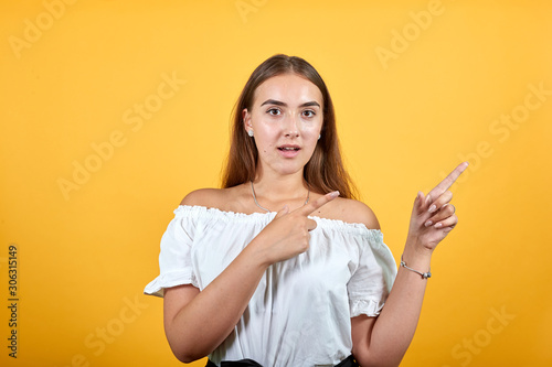 Confused attractive young woman isolated on orange background in studio pointing aside with opened mouth, shocked in casual white shirt. People sincere emotions, lifestyle concept.