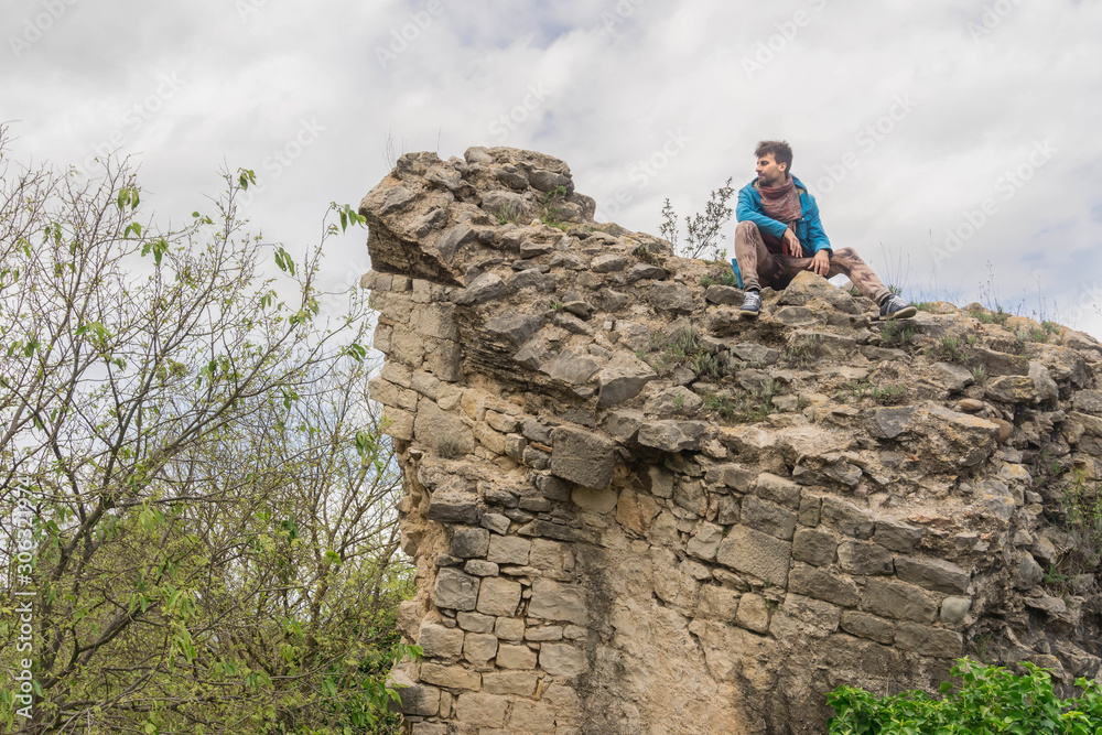 Young man sitting alone on abandoned fortress stone wall with cloudy sky background. Solo traveler concept.