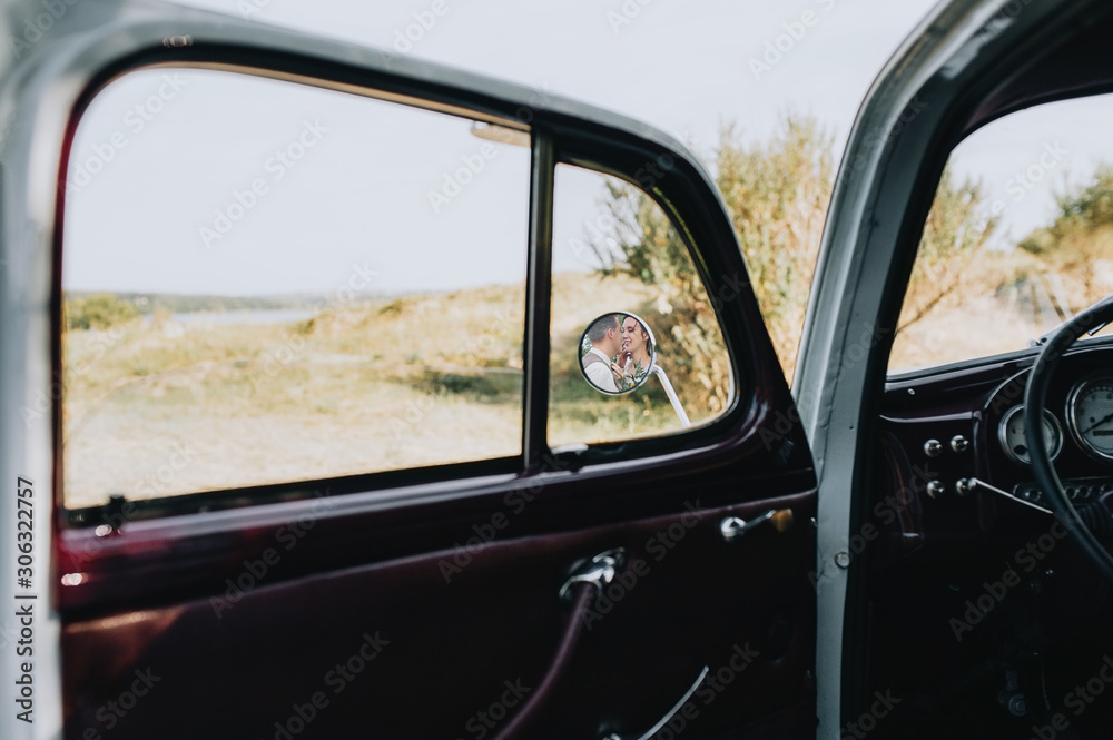 Smiling and in love newlyweds hug, and are reflected in the side mirror of the car in the interior. Wedding portrait of a stylish bride and groom. Photography, concept.
