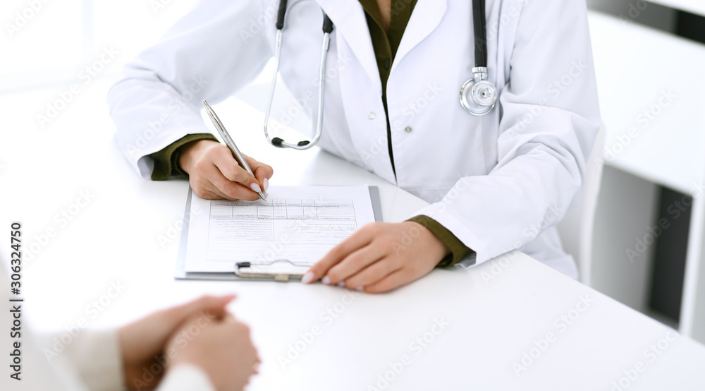 Woman doctor and patient sitting and talking at medical examination at hospital office, close-up. Therapist filling up medication history records. Medicine and healthcare concept Woman doctor and patient sitting and talking at medical examination at hospital office, close-up. Therapist filling up medication history records. Medicine and healthcare concept