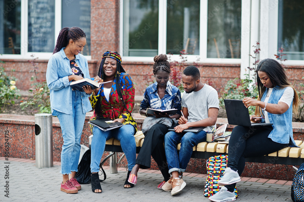 Fototapeta premium Group of five african college students spending time together on campus at university yard. Black afro friends studying at bench with school items, laptops notebooks.