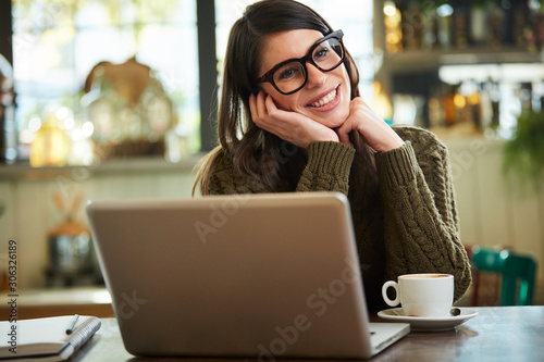 Beautiful caucasian brunette with eyeglasses leaning on table while sitting in cafe. On table is laptop.