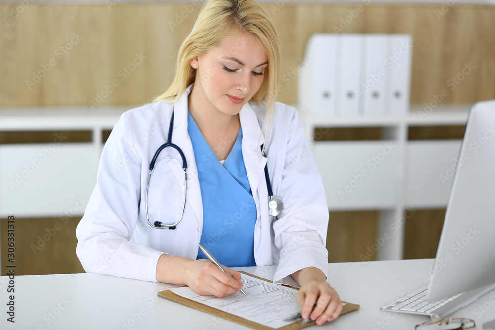 Doctor woman at work while sitting at the desk in hospital or clinic. Blonde cheerful physician filling up medical records form. Data and best service in medicine and healthcare Doctor woman at work while sitting at the desk in hospital or clinic. Blonde cheerful physician filling up medical records form. Data and best service in medicine and healthcare
