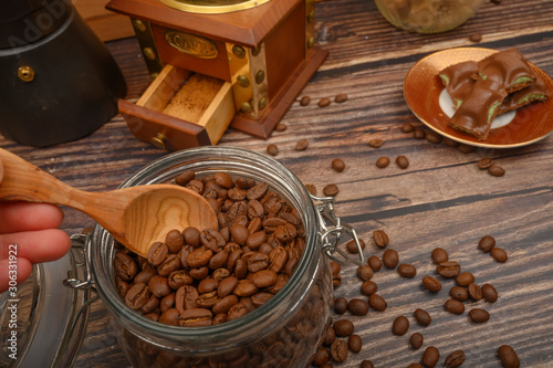 The girl's hand takes a wooden spoon of coffee beans from a glass jar, a coffee grinder, pieces of chocolate on a wooden background. Close up.