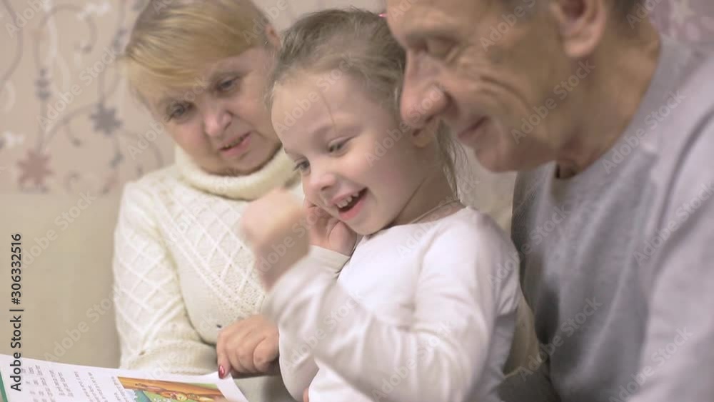 grandfather and grandmother are reading a book with their granddaughter sitting at home on the couch, family values grandfather and grandmother are reading a book with their granddaughter sitting at home on the couch, family values