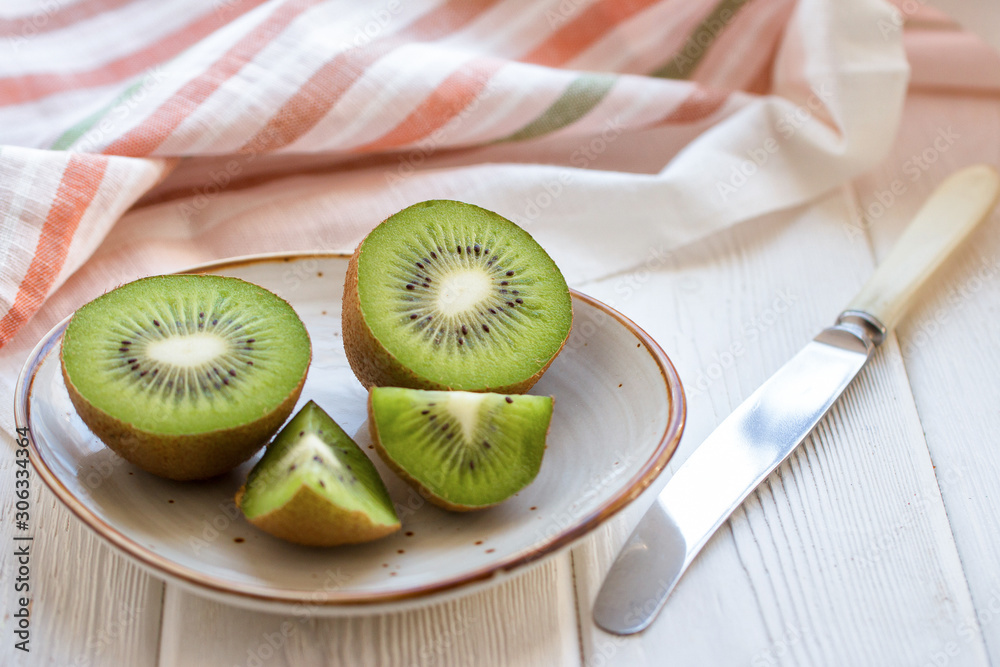 Sliced fresh kiwi fruit on a plate on a light wooden table with a knife and a striped linen napkin. Front view.