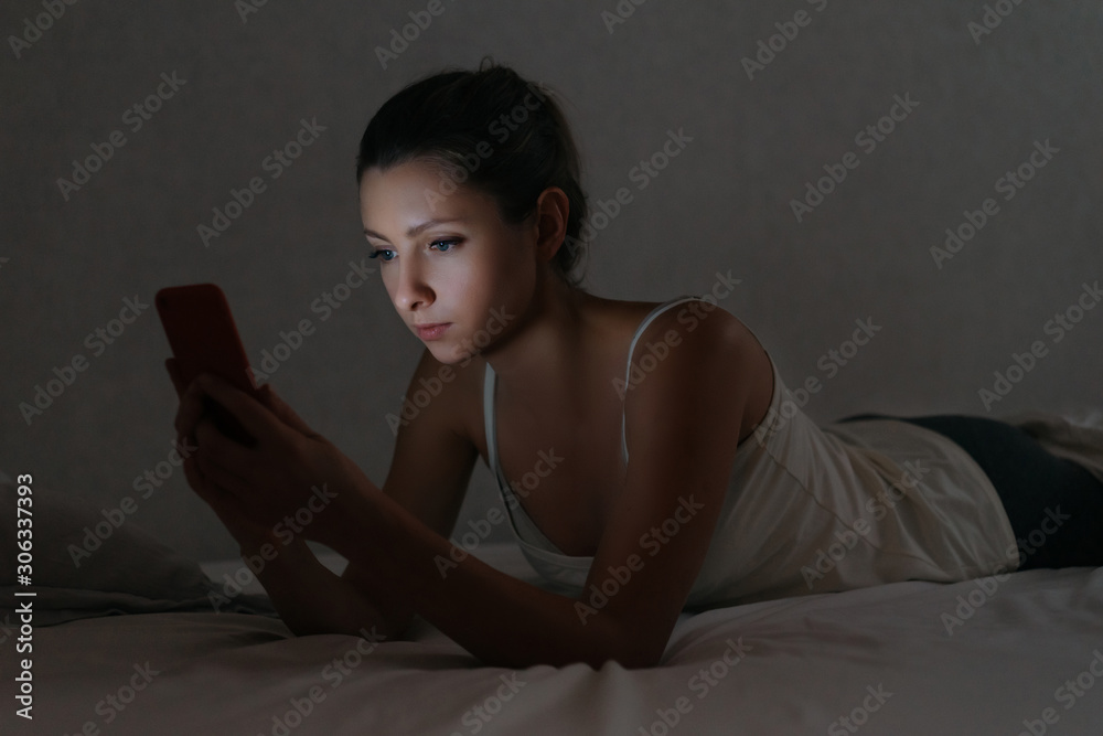 Young woman messaging with smartphone in bed at night. Social network dependence. Gadget addict concept. Harm of using phone before going to bed Young woman messaging with smartphone in bed at night. Social network dependence. Gadget addict concept. Harm of using phone before going to bed