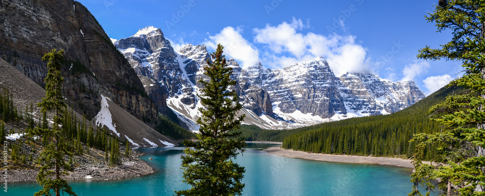This pristine Moraine Lake overlooks the icy rocky mountains and pine forest. The light breeze gently ripples the turquoise water towards the rocky edge on this partially cloudy day.