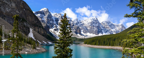 Fototapeta Naklejka Na Ścianę i Meble -  This pristine Moraine Lake overlooks the icy rocky mountains and pine forest. The light breeze gently ripples the turquoise water towards the rocky edge on this partially cloudy day.