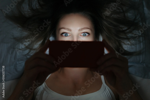 Young female addictively staring at her smartphone late at night in bed. Focused on eyes. Phone dependence concept. Horizontal shot.