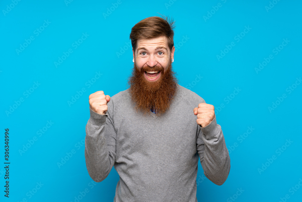 Redhead man with long beard over isolated blue background celebrating a victory in winner position Redhead man with long beard over isolated blue background celebrating a victory in winner position