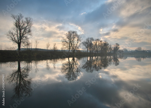 Calm autumn morning landscape with fog and warm sky over the pond surrounded by trees with the beautiful reflections of clouds and trees in the water