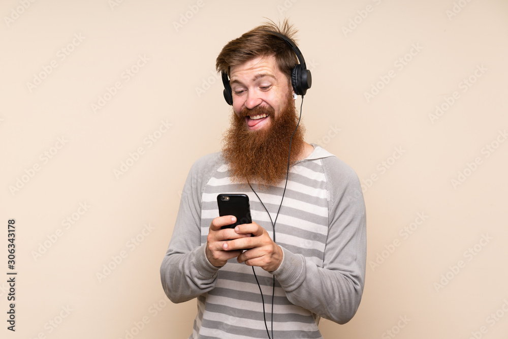 Fototapeta premium Redhead man with long beard over isolated background using the mobile with headphones and singing