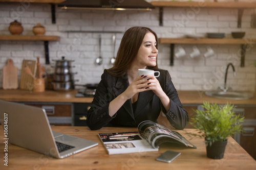 smiling beautiful business woman checking daily news in laptop and reading magazine in the kitchen while drinking morning coffee before going to office . multitasking, rituals photo