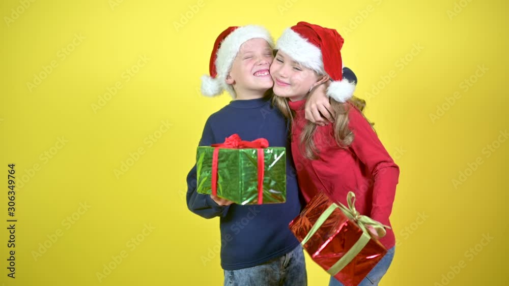 Cute children in santa claus hats give presents to each other and hug, on a yellow studio background. Holidays and fun. Christmas and New Year. Cute children in santa claus hats give presents to each other and hug, on a yellow studio background. Holidays and fun. Christmas and New Year.