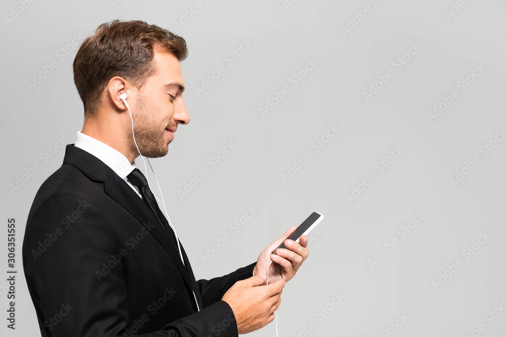 side view of handsome and smiling businessman in suit with earphones using smartphone isolated on grey side view of handsome and smiling businessman in suit with earphones using smartphone isolated on grey