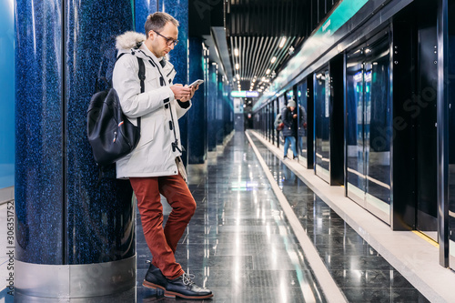 Lonely attractive adult man with glasses in white jacket and in red trousers stands leaning on a column and holding black backpack. Male use smartphone while wait subway train. Mobile phone addiction.