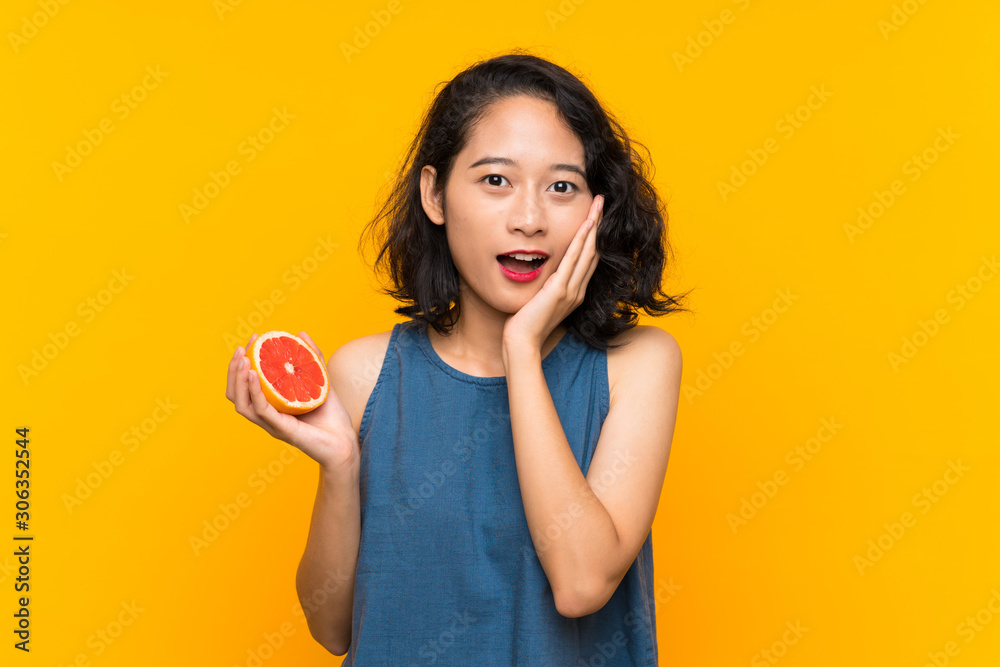 Young asian girl holding a grapefruit over isolated orange background with surprise and shocked facial expression Young asian girl holding a grapefruit over isolated orange background with surprise and shocked facial expression