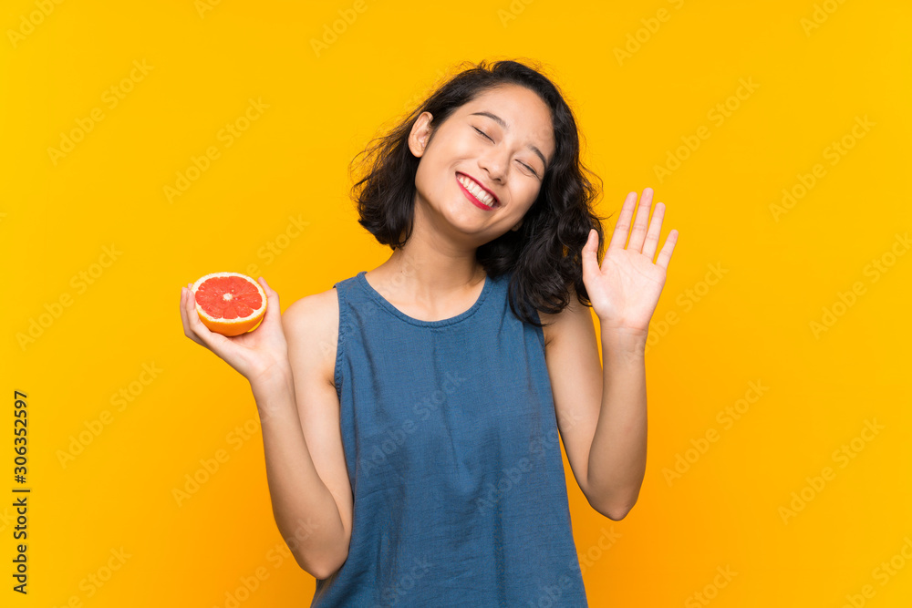 Young asian girl holding a grapefruit over isolated orange background saluting with hand with happy expression Young asian girl holding a grapefruit over isolated orange background saluting with hand with happy expression