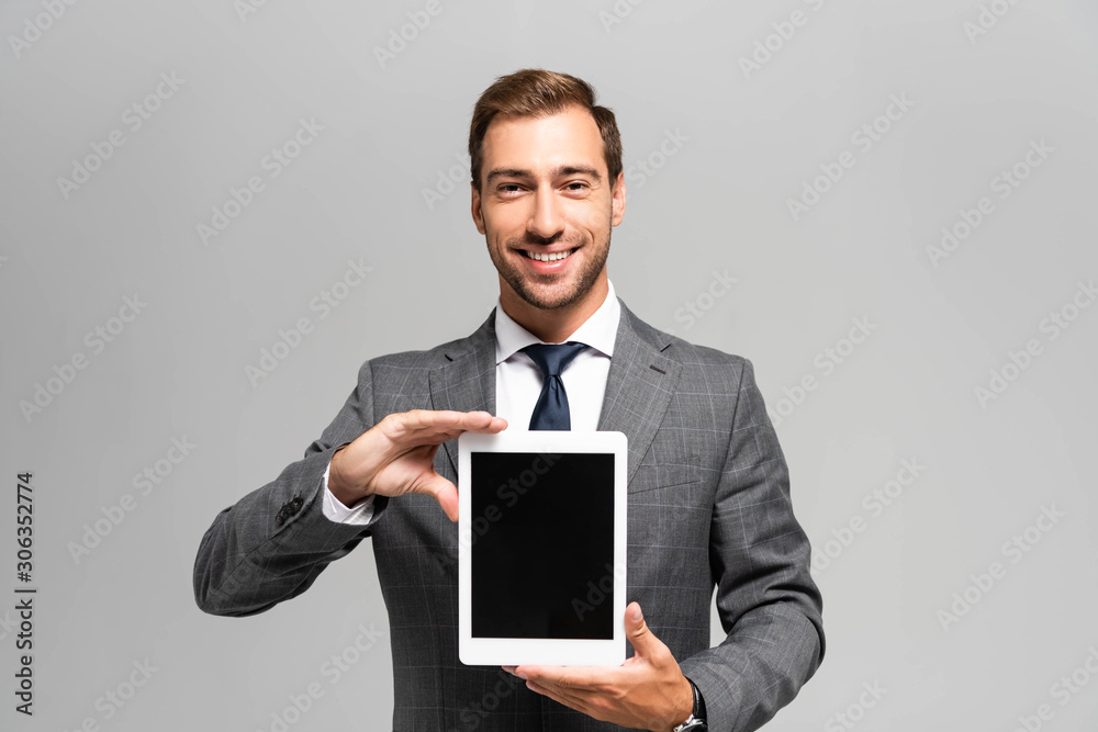 handsome and smiling businessman in suit holding digital tablet isolated on grey handsome and smiling businessman in suit holding digital tablet isolated on grey