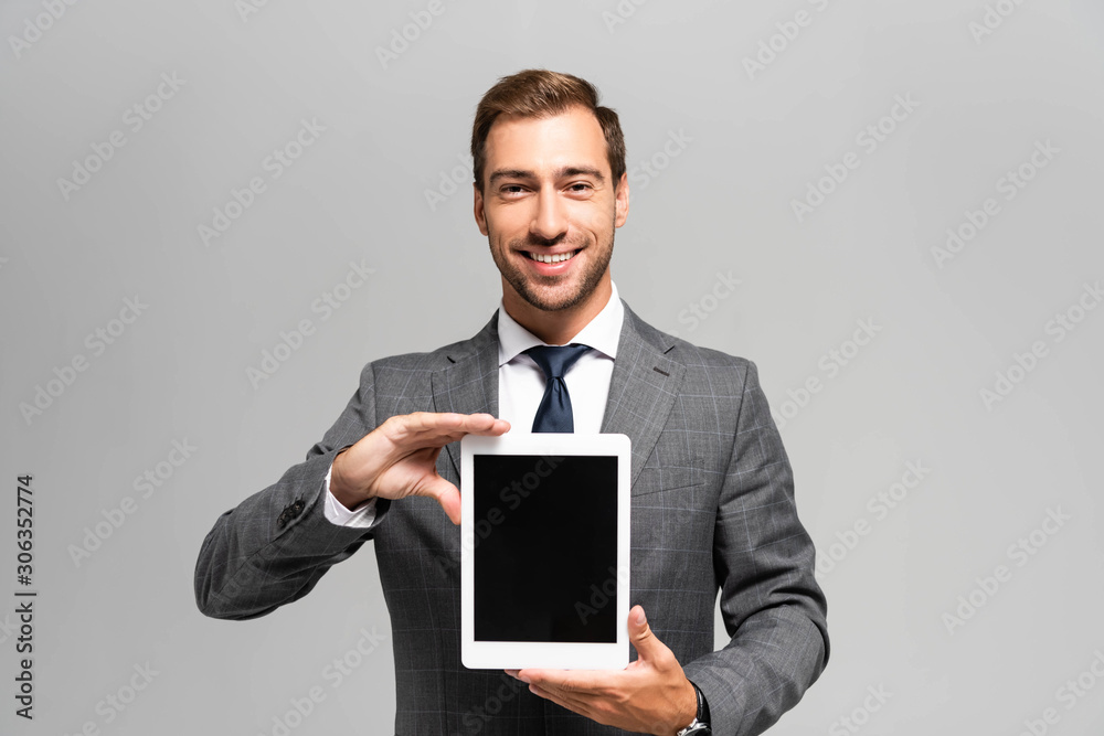 handsome and smiling businessman in suit holding digital tablet isolated on grey