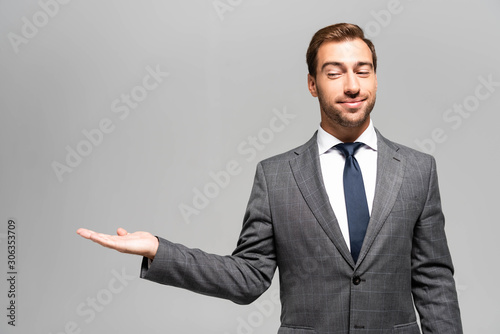 handsome and smiling businessman in suit pointing with hand isolated on grey