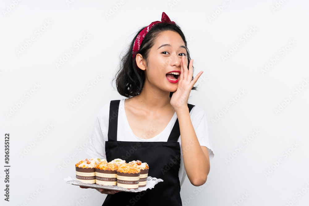 Young asian girl holding lots of muffin cake over isolated white background shouting with mouth wide open Young asian girl holding lots of muffin cake over isolated white background shouting with mouth wide open