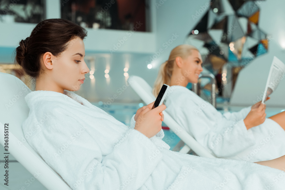 selective focus of attractive woman using smartphone and friend reading newspaper on background in spa selective focus of attractive woman using smartphone and friend reading newspaper on background in spa