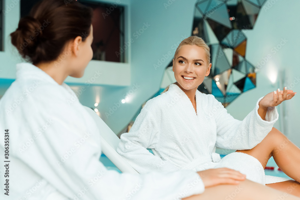 attractive and smiling friends in white bathrobes talking in spa attractive and smiling friends in white bathrobes talking in spa