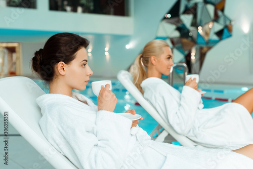 selective focus of attractive woman in white bathrobe holding cup and friend on background in spa