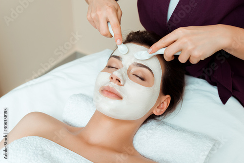 cropped view of cosmetologist applying face mask on attractive woman in spa