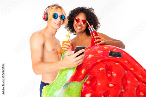 Pretty african female and caucasian blonde man stands in swimsuit with rubber beach mattresses, drinks juice and listen to music isolated on white background